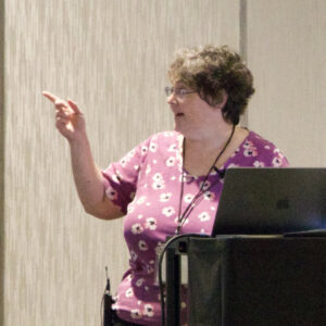 Jill presenting at Macstock 8. Right hand extended, pointing toward presentation on screen.