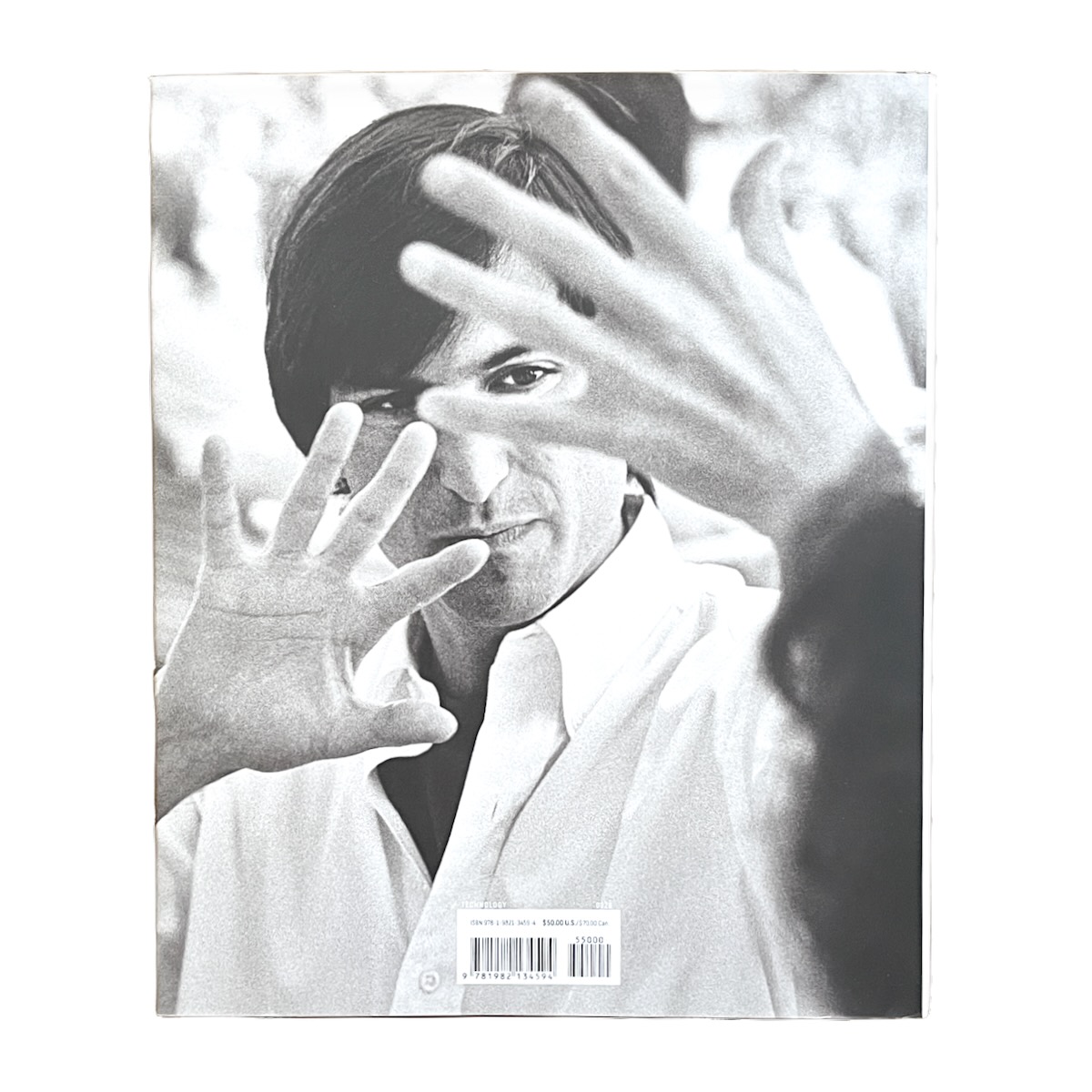 Apple The First 50 Years by David Pogue Back Cover. Black and White photo of Steve Jobs, his hands raised as he appears to share an idea with a colleague.